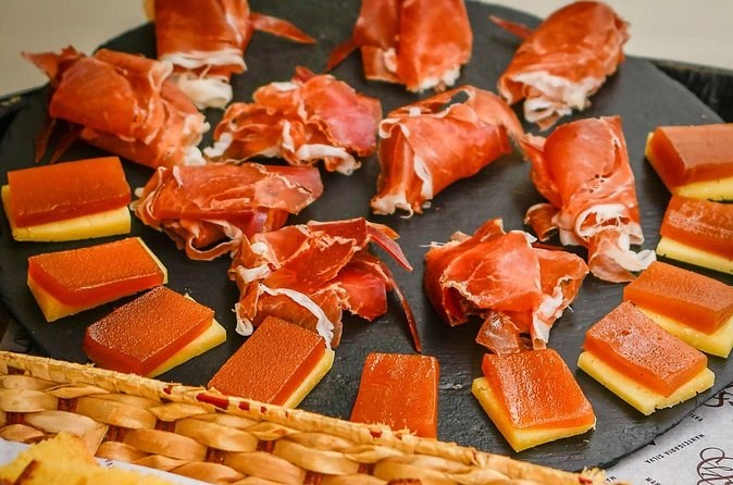 Folded slices of cured ham and squares of cheese topped with quince paste on a black slate board, beside a wicker bread basket